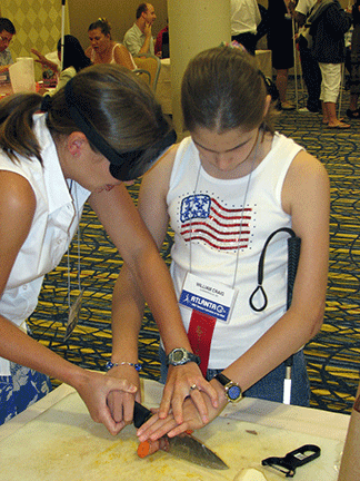 A mentor under sleepshades shows a teenage girl how to slice a carrot.