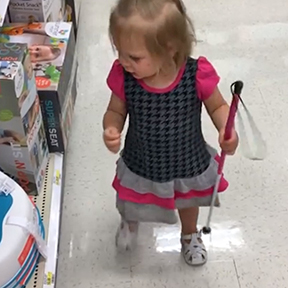 In a store, the child holds her cane in one hand as she looks at items on a shelf.