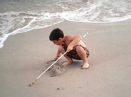 A boy plays in the sand on the beach while holding his cane.