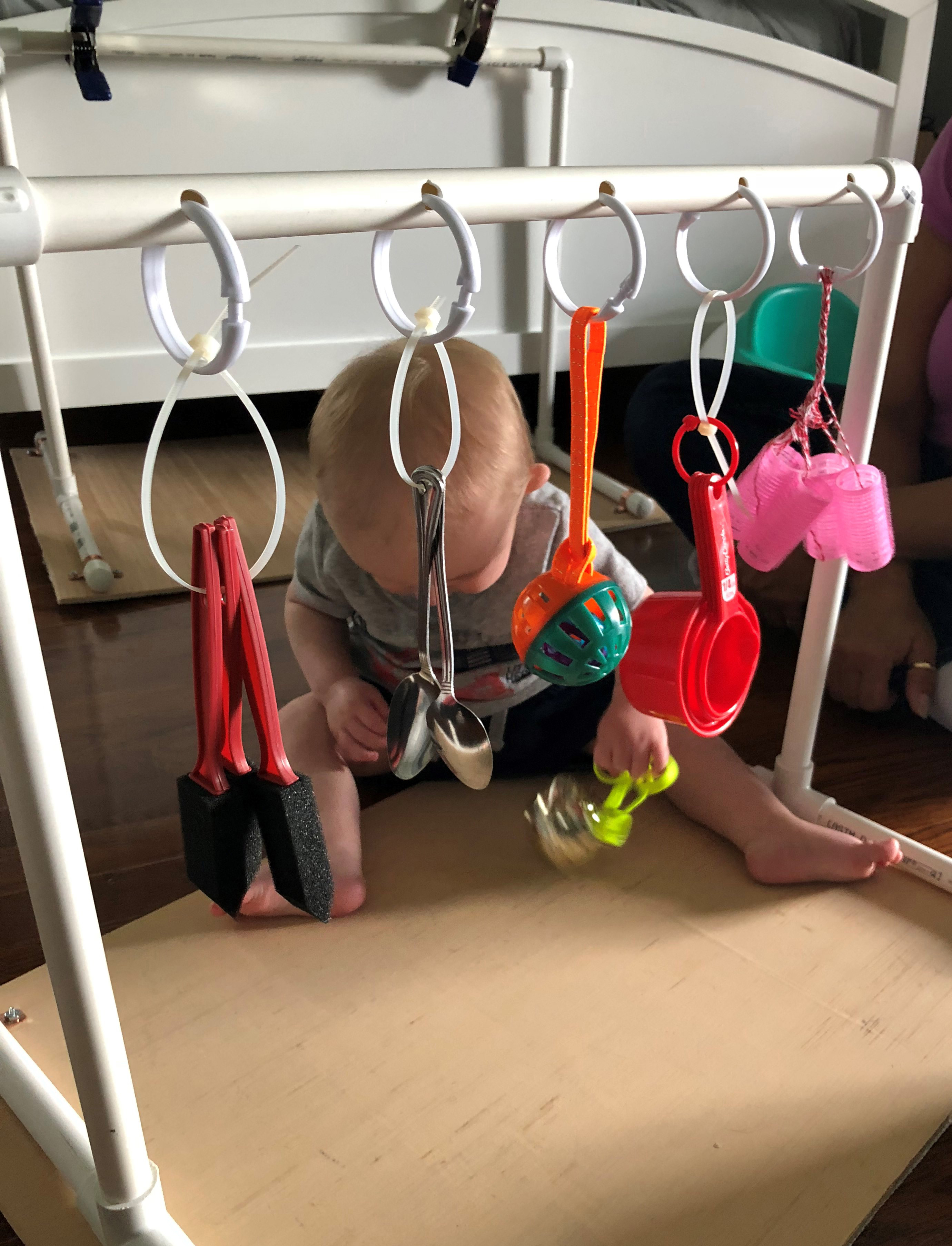 Finn plays on the floor near a mobile with hanging spoons, curlers, paintbrushes, and measuring cups.