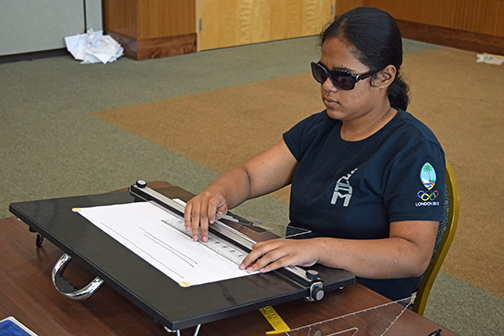 Mara Oneichu sits at a drafting board, using a print/Braille ruler.