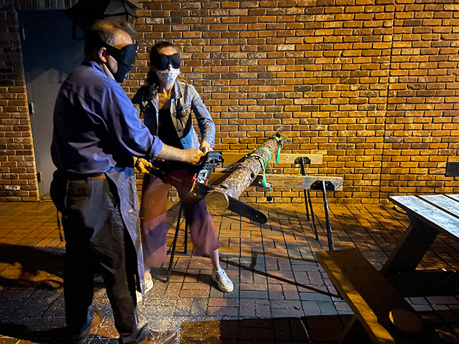 Mark Riccobono helps a teacher wearing sleepshades as she cuts a log with a gas-powered saw.