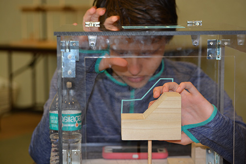 A participant examines a wooden model inside a plexiglass box. The box has tactile diagrams showing each side of the object, teaching the concept of a multi-view drawing.