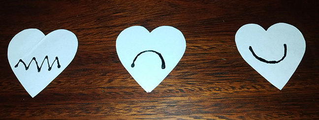 On a wooden background there are three hearts made with white cardboard. The first heart, located on the left side, has zig-zag lines. The middle heart contains a semicircle with the concave part facing downward. The heart located on the right side contains a semicircle with the concave part facing upward. The designs on these hearts were made with black embossed glue.