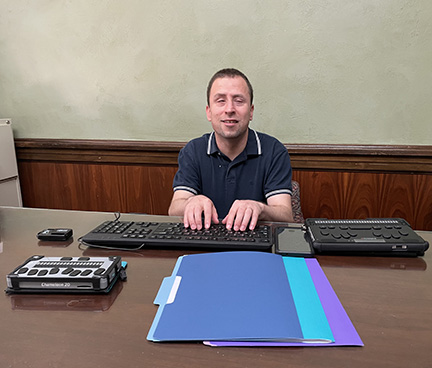 Chuy Vaca sits at a table with a BrailleSense, a Chameleon, an iBill, a cell phone, and a keyboard.