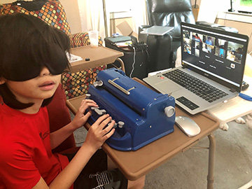 A young boy wearing learning shades sits in front of a computer showing a Zoom session and working on a Brailler.