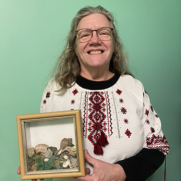 Lydia Schuck wears a vyshyvanka, a traditional Ukrainian embroidered blouse. She holds a shadowbox containing sea glass and Ukrainian coins. 
