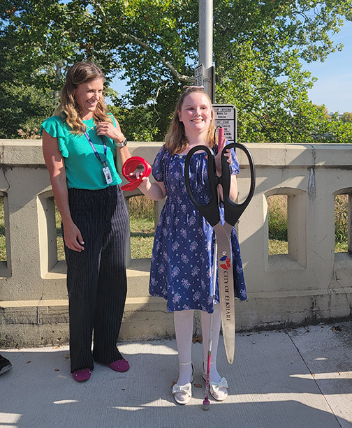 Willow Townsend stands in front of a pedestrian signal pole, holding a roll of red ribbon and a huge pair of scissors labeled &ldquo;City of Elkhart.&rdquo;