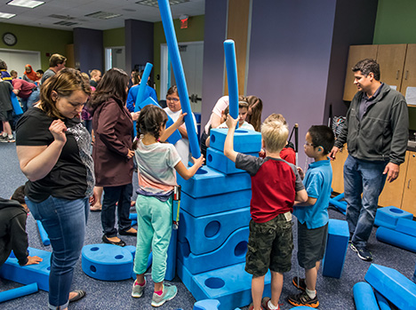 A group of children work together to build a tall structure using large blue foam blocks and tubes in a classroom-like space, while several adults stand nearby observing and assisting.