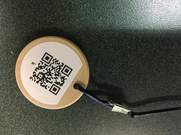 Close-up picture of a wooden coin with a QR code
label attached to it. A leather lanyard is attached to the coin
through a hole drilled in it. The other end of the lanyard is
similarly attached to the 3-D printed point.
