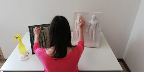Person sitting, viewed from behind. With the left hand, she touches her clay copy of bas-relief and with the right one the original in alabastrine plaster.
