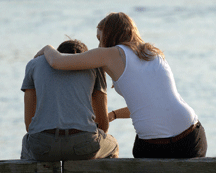 A man and woman sit with their backs to us. The woman has her arm around the man’s back and appears to be comforting him.