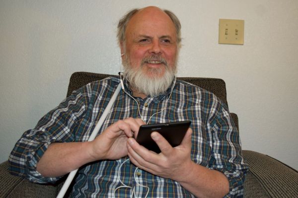 A man sits in a chair holding an iPad and listens to NFB-NEWSLINE with headphones.
