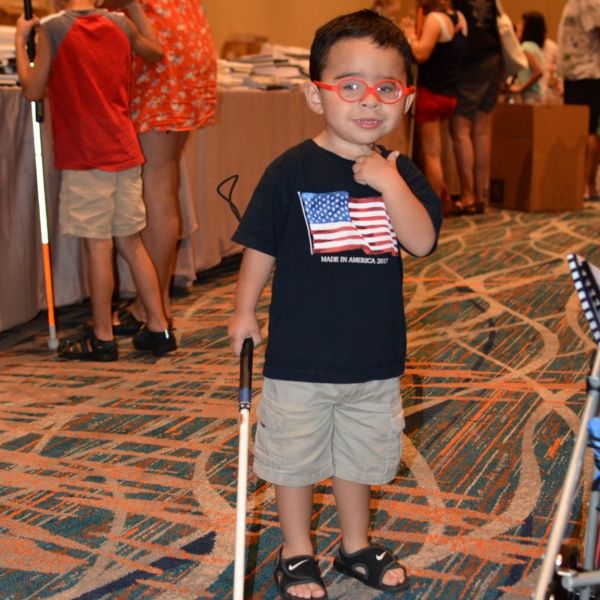 A young boy holds a white cane and gives the thumbs up sign.