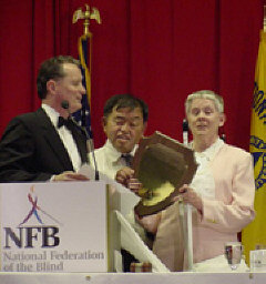 President Maurer speaks at the podium while Al and Sharon Maneki hold their plaque.