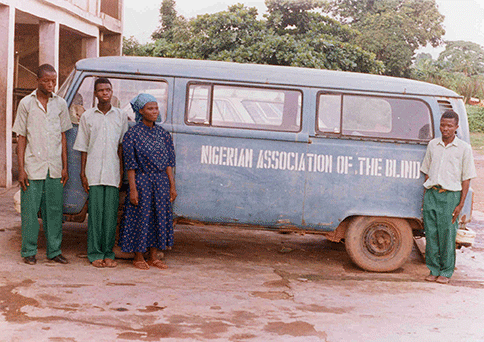 A group of blind Nigerians stands beside a bus. On the side of the bus is the name Nigerian Association of the Blind.