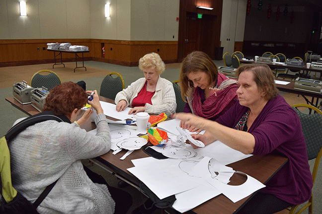 Participants cutting out materials during a hands-on activity: (Left to right) Mary Jo Partyka (NJ) cuts out a face that has raised lines on its perimeter for accessibility while Rebecca Sherman (ID) reads the lesson plan for this activity from the NFB BELL Academy curriculum. Alison Steven (ID) and Debbie Wunder (MO) tactually review their cut-out faces.