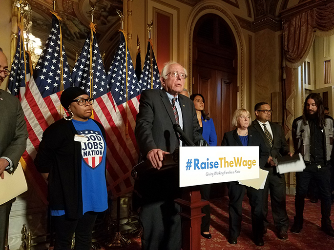 Senator Bernie Sanders stands at a podium with a #RaiseTheWage banner to announce the new bill.