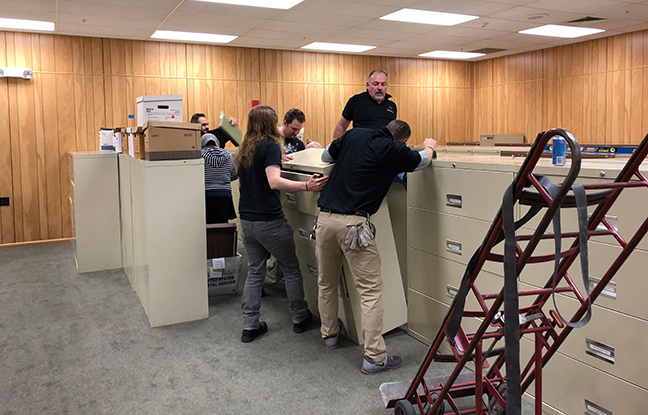 A group of staff wrestle with a filing cabinet to get it into its proper place.