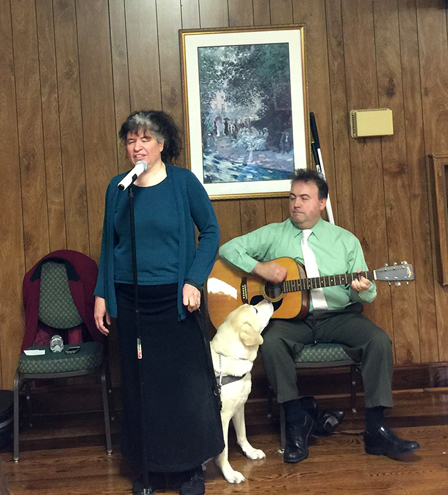 Melissa Riccobono stands and sings into a microphone. Mark Riccobono sits behind her playing the guitar. Melissa’s guide dog sniffs at Mark’s guitar.