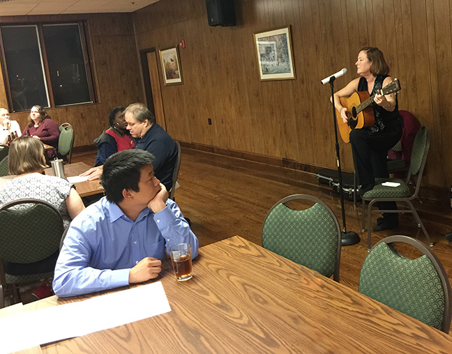 Carla McQuillan plays her guitar and sings into the microphone in front of a group of people who sit listening.