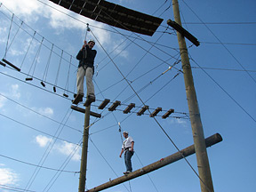 Two men from the Utah mentoring program take part in a high ropes course event. One walks on a rope bridge and the other climbs a horizontal utility pole.