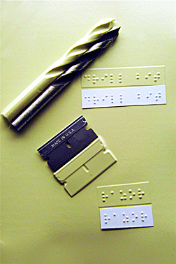 Sample: A sharp drill bit and a razor blade lie beside their identical thermoformed&nbsp;shapes with identifying thermoformed braille labels. 