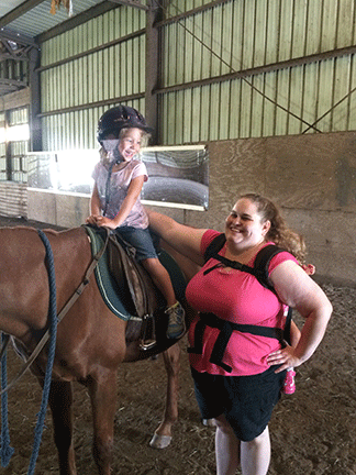 While carrying a child in a backpack, Brandy Wojcik encourages a little girl who is seated on a horse.