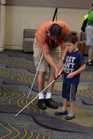 A man using a long white cane teaches cane technique to a young boy.