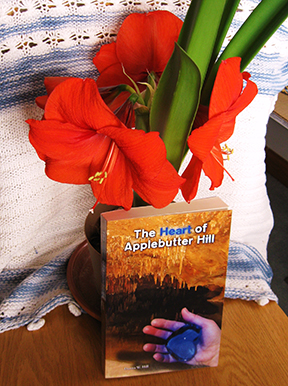 The cover of The Heart of Applebutter Hill, showing a hand holding a blue heart, a cave with stalactites in the background. The book leans against a flowerpot with a beautiful amaryllis.