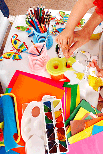 Surrounded by crafts materials, the hands of a child and an adult dip a paintbrush into water-based paint.