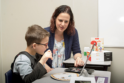 Using Osmo tiles, a boy adds toppings to a fake pizza as his teacher looks on.