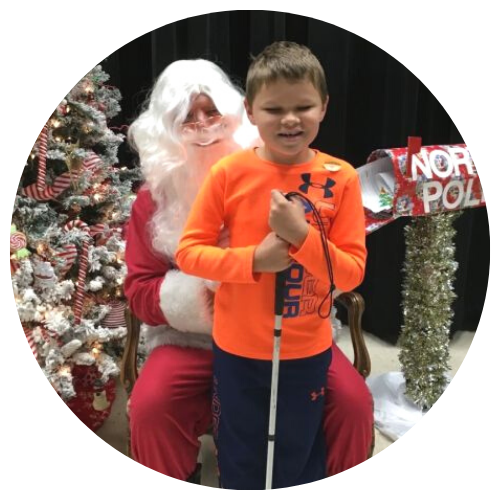A young boy with a white cane sits on Santa's lap, Christmas tree and north pole mailbox in the bachground