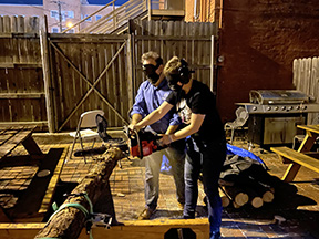 Mark Riccobono helps a participant use the chainsaw to cut a long log. They both wear sleepshades and masks.