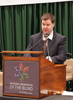 Justin Young, a white male wearing a gray suit, white shirt, and multi-colored tie, speaking at the podium during the 2022 National Federation of the Blind National Convention.