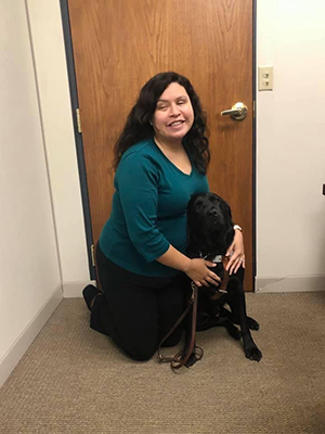 Danielle McCann, wearing a green shirt and black pants, kneeling with her black Labrador Retriever retired guide dog Schulz.