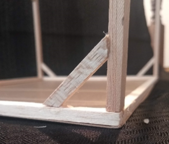 A closeup of a braced corner of a partially constructed balsa model. The trapezoidal-shaped brace, the corner column, and the base of the model form an isosceles right triangle.