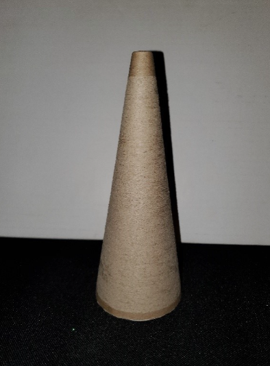 The cone has a height of 7” and a 3” diameter circular base. It is a right circular cone, so the vertex of the cone is positioned directly above the center of the base. About ½” of material has been removed from the vertex of the cone, creating a smaller circular apex rimmed by the cut edge. The cone is positioned on a tabletop. This photo shows the snipped cone in elevation: The base of the cone sits flat on the table and thus is not visible per se; the long, curved surface of the cone tapers upwards, with the cut vertex at the top. The point of view is face-on to the cone.