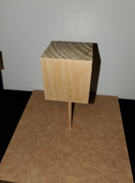 A 3” wood cube mounted above a rectangular base using a dowel. The base is a rigid, wood platform. The cube is mounted on the vertical dowel at an angle so that none of its faces align with the base.
