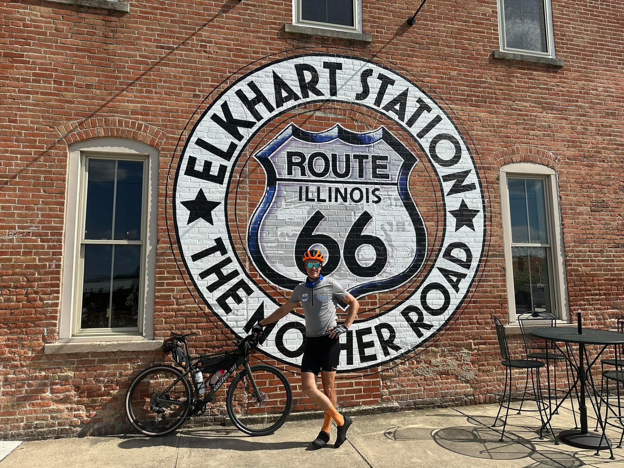 Dan stands with bike in front of a brick building displaying a large Route 66 sign in Illinois