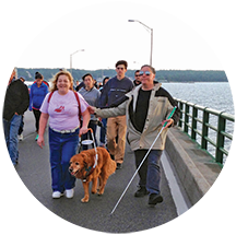 Two blind adults walk across a bridge one uses a guide dog, the other a cane.