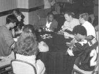 Seven people sit at a round table with cards and chips visible on it.