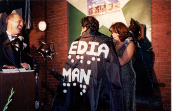 A man stands with his back to the camera while Betsy Zaborowski ties his cape on.