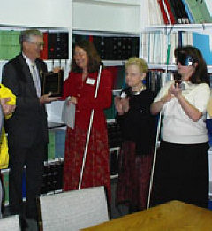 Julie Deden presents a plaque to Rod Sylvan while Diane McGeorge and Jamie Foster look on.   Behind them we see many shelves of Braille books.
