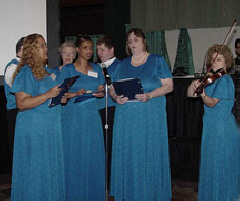 A group of singers, the women in long blue dresses, stand singing with a violinist beside them.