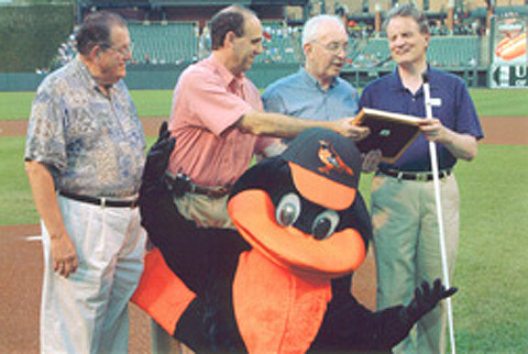The Robert Dole Award presentation took place on the field shortly before the Orioles game began.  The Orioles mascot waves at the camera during the awards ceremony.   Left to right the participants are Al Ruppersberger, award chair; Dr. Michael Weinrigh, chair of the Volunteers for Medical Engineering, Inc.., board of directors; John Staehlin, founder and president of VME; and NFB President Marc Maurer.