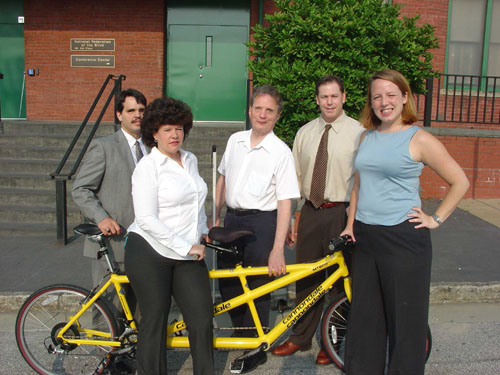 NFB staff members taking part in the Face of America ride stand with President Maurer in front of the National Center.   Shown are Maurice Peret, Holly Mooney, Marc Maurer, John Brennan, and Karen Friedman.  The tandem bicycle ridden by the Mooney-Peret team is visible behind Mrs. Mooney.