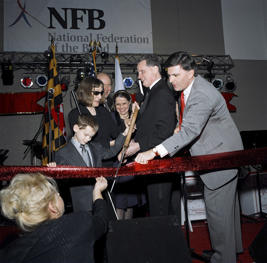 Near the close of the grand opening program, representatives of the organizations making million-dollar gifts to the capital campaign joined dignitaries and Federation representatives on stage for the ribbon-cutting ceremony.   Here President Maurer uses oversized scissors to cut the broad textured ribbon.   Pictured left to right are Jason Polanski, a seven-year-old from Maryland representing the next generation of blind people; Barbara Walker Loos, president of the American Action Fund for Blind Children and Adults; Wally O'Dell, chairman and CEO of Diebold, Inc.; Mary Ellen Jernigan, NFB executive director of operations; (almost hidden) Steve Marriott, senior vice-president of culture and special events for Marriott International, Inc.; Marc Maurer, president of the National Federation of the Blind; and Robert L. Ehrlich, Jr., governor of the state of Maryland.