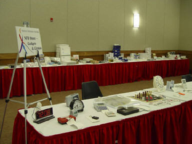 Pictured here before the crowd arrived is the NFB Store's popular display, Gadgets and Gismos.  On the front table are items that many newly blind people know nothing about: talking clocks and calculators, raised-dot and  large-print timers, games, and sewing aids. Behind is the stock table to replenish supplies as items are sold.