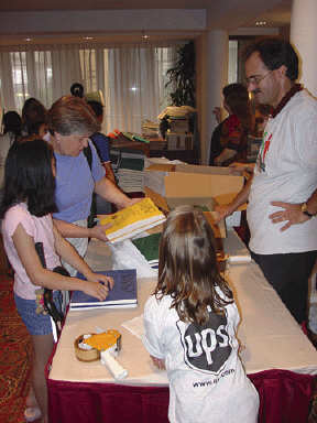 At the left a girl of about eleven is reading a Braille book while standing on one side of a table being used to pack up books.  Her mother holds another Braille book and talks to her.   At the center of the picture a little girl leans across the table from the UPS side.   Her back is to the camera and her T-shirt has a large UPS logo on it.   A tape gun can be seen beside her.   A UPS volunteer stands across the table facing the Braille reader.